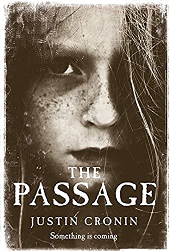 A book cover for 'The Passage' by Justin Cronin. The cover is sepia-toned and features a close-up image of a young girl's face looking directly at the viewer, with strands of her hair partially covering her face. Her expression is intense and haunting. There are speckles and spots across the image, giving it a gritty, aged appearance. The title 'The Passage' is displayed in bold, white lettering at the top, and the author's name 'Justin Cronin' is just below it in smaller print. At the bottom, there's a tagline in a smaller font that reads 'Something is coming,' adding an ominous tone to the overall design.